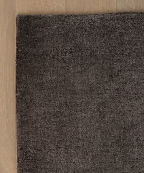 A corner of the dark brown Bristol Rug rests on a light wooden floor, showcasing the texture contrast between the soft rug and the smooth wood surface.