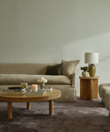A minimalist living room with a beige sofa, brown cushion, round wooden coffee table with books, flowers, and a candle, plus a side table with lamp and vase—all set on the Bristol Rug for added warmth and character.
