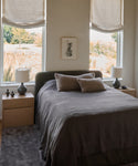 A cozy bedroom features a neatly made bed with a gray blanket, beige pillows, wooden nightstands and matching lamps, plus the Bristol Rug underfoot. Large windows with fabric shades fill the room with natural light.