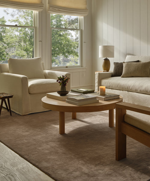 A cozy living room with a cream armchair, beige sofa, wooden coffee table, side lamp, large windows, and the Bristol Rug—a handwoven piece made in India in neutral tones—adds warmth and style to the space.