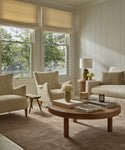 Bright living room with large windows, cream armchairs, a white sofa, round wooden coffee table with books and flowers, side table with a lamp, neutral décor, the Bristol Rug handmade in India, and soft natural light.