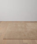 The Bristol Rug, in beige, is handwoven and placed on a light wooden floor in a minimalist room with a plain white wall backdrop.
