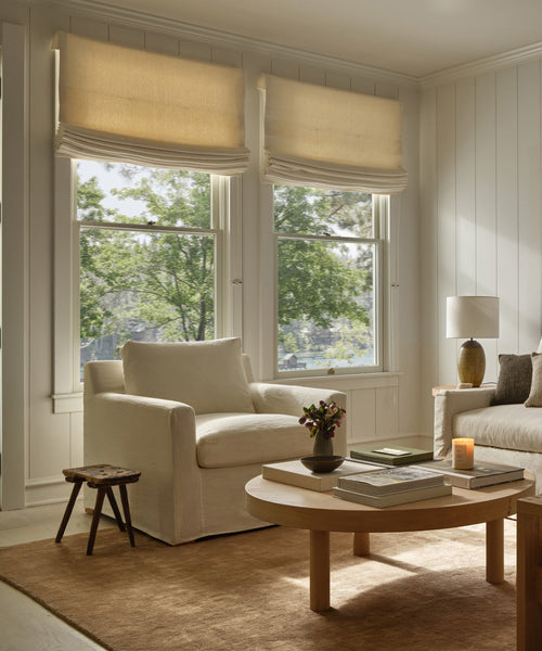 A bright, cozy living room featuring two large windows with beige Roman shades, a white armchair, a beige sofa, a small stool, a side table with lamp, and a Rug Swatch in mocha tones beneath a round wooden coffee table.