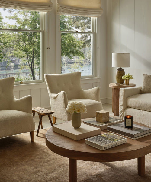 Bright living room with cream armchairs, a matching sofa, and a round wooden coffee table topped with books, flowers, and a candle. A Rug Swatch in Mocha enhances the space, while large windows with Roman shades offer views of green trees.