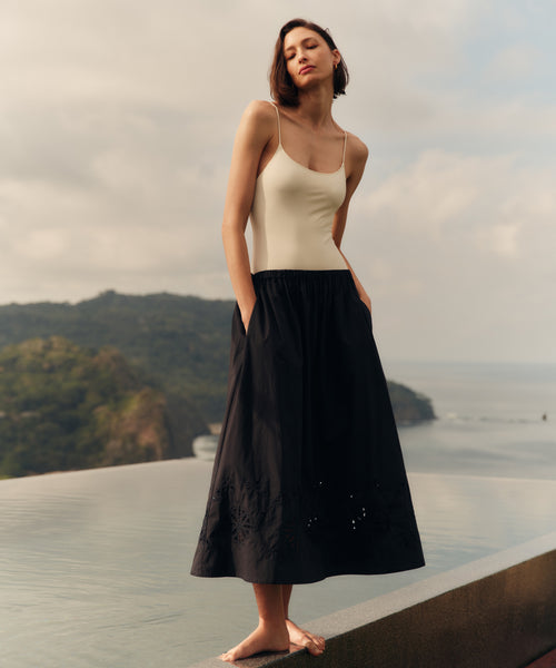 Woman wearing a cream top and black skirt standing on a rooftop with a scenic view of mountains and sea.