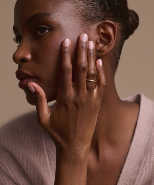 A woman with short hair gently touches her face, displaying natural nails and the Brook Gold Band. She wears a soft light pink sweater and gazes thoughtfully to the side, set against a neutral backdrop.