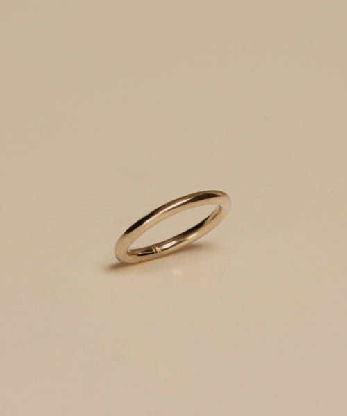 The Brook Gold Band, a sleek and thin 14k gold ring, stands upright on a smooth beige surface, casting a subtle shadow below it.