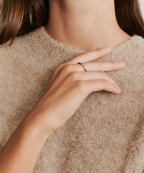 A person wearing the Brook Gold Band, a simple 14k solid gold ring, rests their hand near their chin while dressed in a fuzzy beige top.