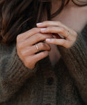 A person wearing a fuzzy brown cardigan gently touches their fingers, showcasing the Brook Gold Band—a simple 14k gold ring. Their long brown hair peeks into view, enhancing the elegant, timeless heirloom style.