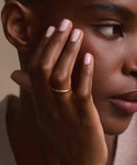 A close-up of a woman's face and hand, highlighting her natural makeup, light pink nails, and the Brook Gold Band on her finger as she gazes calmly to the side.