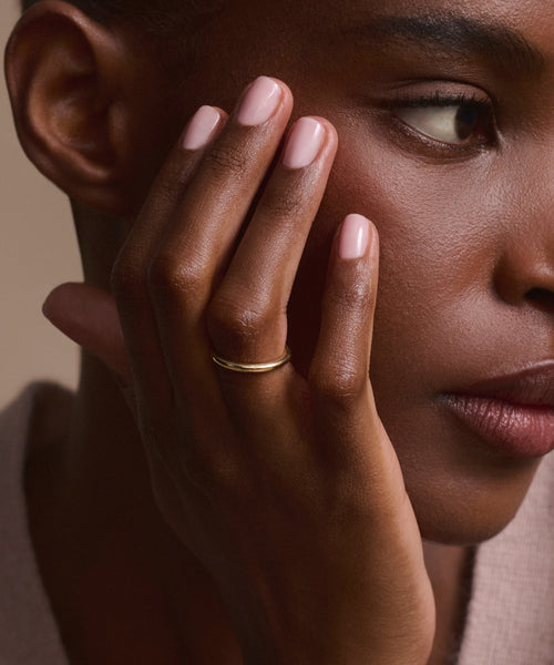 A close-up of a woman's face and hand, highlighting her natural makeup, light pink nails, and the Brook Gold Band on her finger as she gazes calmly to the side.