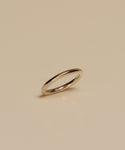 The Brook Gold Band, a simple and thin 14k gold ring, stands upright on a neutral beige surface, casting a faint shadow beneath it.