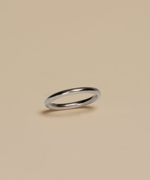 The Brook Gold Band, a slim white gold ring, rests on a plain beige surface with a subtle shadow beneath.