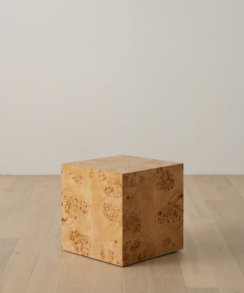 The Sur Side Table is a square wooden cube with a light, natural finish and visible wood grain, evoking a vintage look. It sits on a light wood floor against a plain white wall.