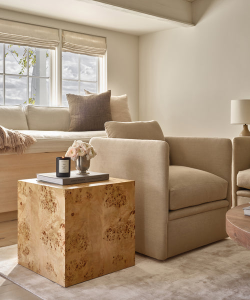 A cozy living room with a beige armchair, the Sur Side Table topped with a candle and flowers, a window seat with pillows, and soft natural light streaming through large windows.