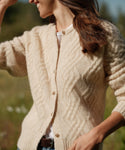 A woman outdoors wears the Cable Crewneck Cardigan in cream over a white top, her brown hair loose and one hand raised near her head.