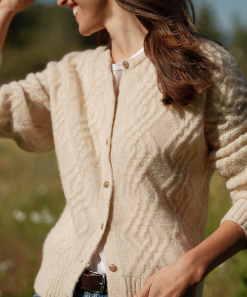 A woman outdoors wears the Cable Crewneck Cardigan in cream over a white top, her brown hair loose and one hand raised near her head.