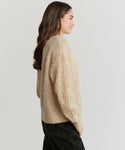 A woman with long brown hair stands in profile against a plain light background, wearing the Cable Crewneck Cardigan in beige and dark jeans.