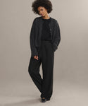 A person stands against a neutral background wearing the Cable Juliette Cardigan with black wide-leg trousers, a black top, and black shoes; one hand rests in a pocket and they have a relaxed expression.