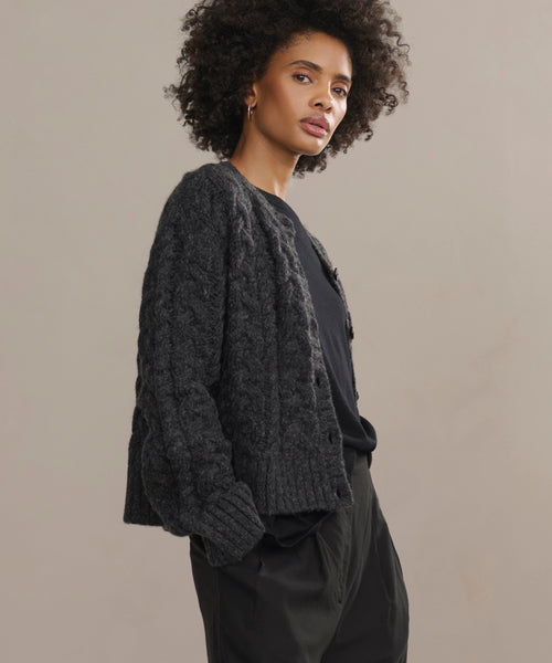 A woman with curly hair stands sideways against a neutral background, wearing the dark grey Cable Juliette Cardigan over a black shirt and dark pants, her hands in her pockets.