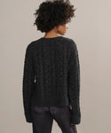 Seen from behind, a person with curly hair wears the Cable Juliette Cardigan in textured dark gray with dark jeans, set against a plain, light-colored background.