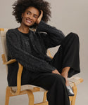 A woman with curly hair smiles while sitting sideways on a wooden chair, resting her head on her hand. She wears the Cable Juliette Cardigan with black pants and shoes, set against a neutral background.