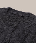 Close-up of the Cable Juliette Cardigan, a dark gray textured knit sweater made from Peruvian yarn, featuring two black buttons and a black fabric label at the neckline, set against a plain light beige background.