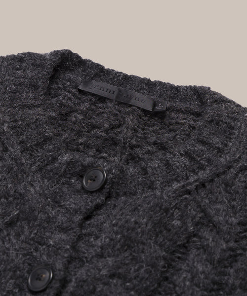 Close-up of the Cable Juliette Cardigan, a dark gray textured knit sweater made from Peruvian yarn, featuring two black buttons and a black fabric label at the neckline, set against a plain light beige background.