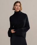 A woman with chin-length brown hair models the black Cable Nell Turtleneck sweater, featuring wide sleeves, paired with a pleated black skirt against a plain light beige background.