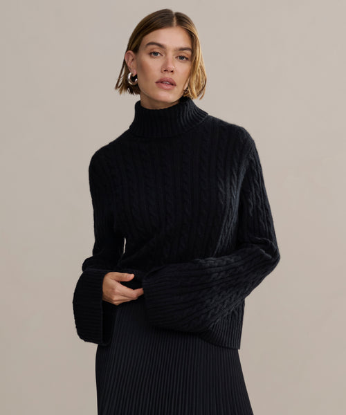 A woman with chin-length brown hair models the black Cable Nell Turtleneck sweater, featuring wide sleeves, paired with a pleated black skirt against a plain light beige background.