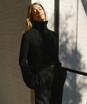 Wearing the Cable Nell Turtleneck and dark trousers, a woman stands by a sunlit wall with her eyes closed, enjoying the sunlight as window shadows create patterns beside her.