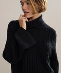 A woman with short blonde hair wears the Cable Nell Turtleneck, a black cable knit sweater with a thick turtleneck, as she looks down and gently touches her face against a plain, neutral background.