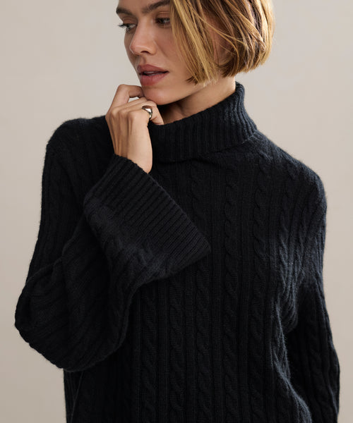 A woman with short blonde hair wears the Cable Nell Turtleneck, a black cable knit sweater with a thick turtleneck, as she looks down and gently touches her face against a plain, neutral background.