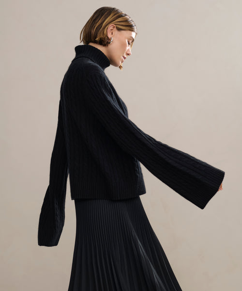 A woman with short, light brown hair wears the black Cable Nell Turtleneck and a black pleated skirt, standing in profile against a neutral background.