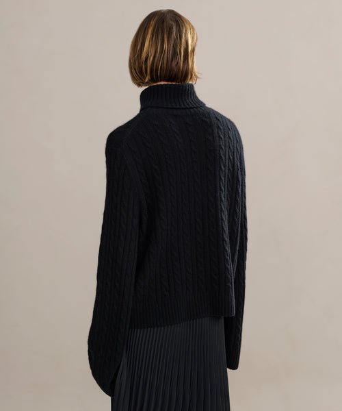 A person with short, light brown hair is shown from behind wearing the Cable Nell Turtleneck and a pleated black skirt against a neutral background.