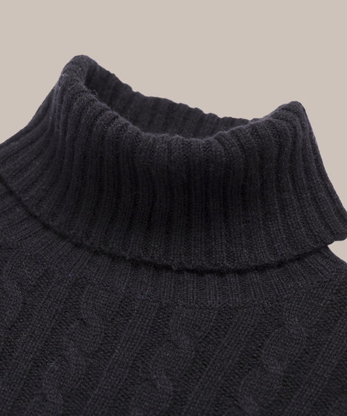Close-up of the Cable Nell Turtleneck in black, featuring a cable knit design and ribbed texture, displayed against a plain beige background.