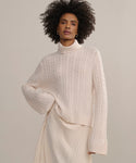 A woman with curly hair stands against a neutral background, wearing the Cable Nell Turtleneck sweater with a boxy silhouette and an ivory pleated skirt.