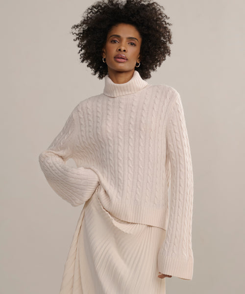 A woman with curly hair stands against a neutral background, wearing the Cable Nell Turtleneck sweater with a boxy silhouette and an ivory pleated skirt.