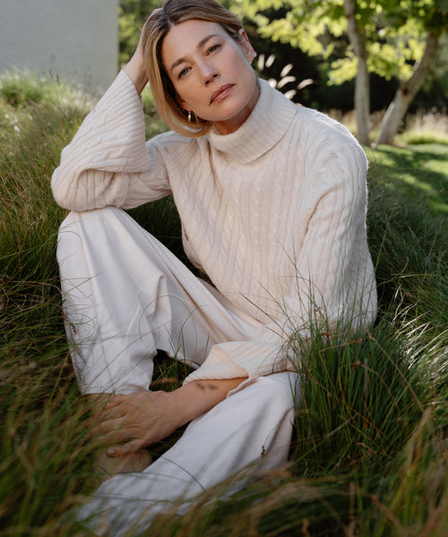 A woman in the Cable Nell Turtleneck ivory sweater with a boxy silhouette and matching pants sits in tall grass, sunlight filtering behind her, as she rests her head on her hand and gazes thoughtfully at the camera.