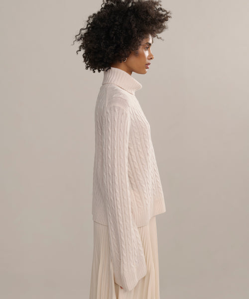 A woman with curly hair stands in profile against a plain background, wearing the Cable Nell Turtleneck—a boxy ivory cable knit sweater—paired with a matching pleated skirt.