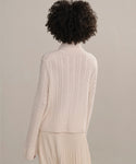 A person with curly hair is shown from behind, wearing the Cable Nell Turtleneck and a pleated skirt against a neutral background.
