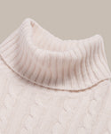 Close-up of the Cable Nell Turtleneck in ivory, showcasing its ribbed collar and textured cable knit pattern against a plain backdrop.