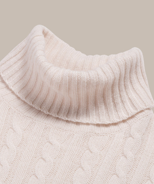 Close-up of the Cable Nell Turtleneck in ivory, showcasing its ribbed collar and textured cable knit pattern against a plain backdrop.