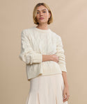 A woman with short blonde hair wears the Cable Serena Crewneck in ivory and a light pleated skirt, standing against a beige background with a neutral expression.