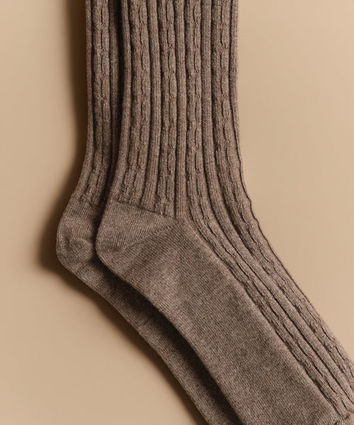 A close-up of Cable Socks in brown, featuring an ultra-stretchy ribbed knit pattern, laid flat on a beige background.