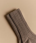 A pair of Cable Socks, ultra-stretchy and ribbed in brown wool, is neatly placed on a beige background.