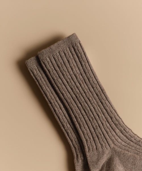 A pair of Cable Socks, ultra-stretchy and ribbed in brown wool, is neatly placed on a beige background.