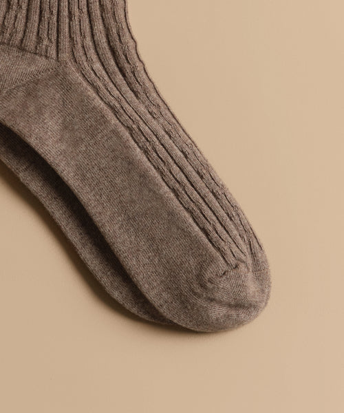 A close-up of a single Cable Sock on a beige background, showcasing its mid-weight knit pattern and ultra-stretchy, comfortable texture.