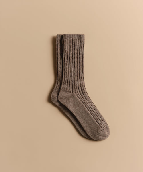 A pair of Cable Socks in ultra-stretchy, ribbed brown wool is neatly stacked on a light beige background.