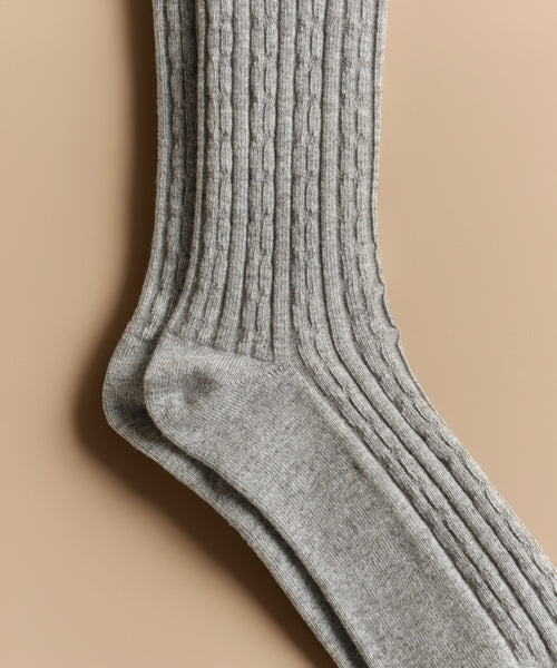 A close-up of two Cable Socks in light gray, laid flat on a beige surface, highlights their textured vertical patterns and soft, ultra-stretchy fabric—ideal for everyday wear.